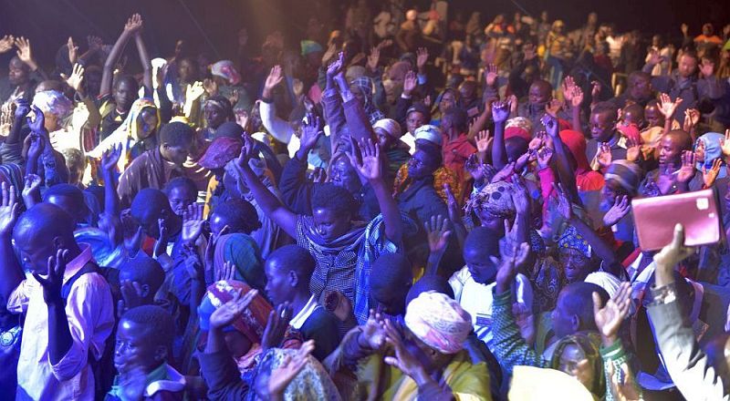 A sea of praying African people with upraised hands, bathed in blue and yellow light