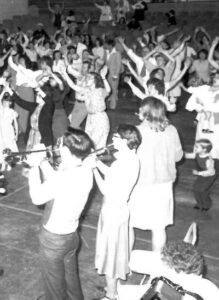 Photo of a 1970s Harvest City worship service; many dancing with raised hands and the orchestra's horn section in the foreground
