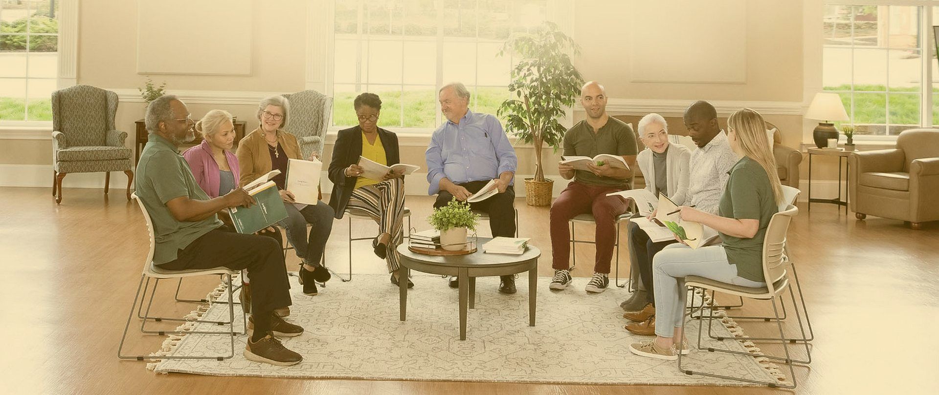 A diverse group of people sit together at a GriefShare support group. They are holding workbooks and warmly interacting.